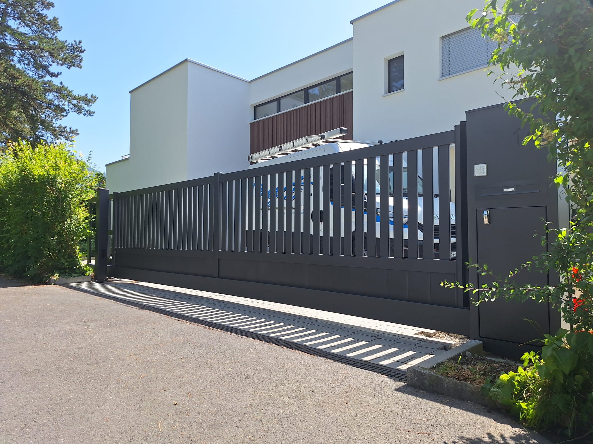 Portail coulissant noir installé devant maison suisse moderne, motorisation fiable, spécialistes portails, environnement verdoyant.