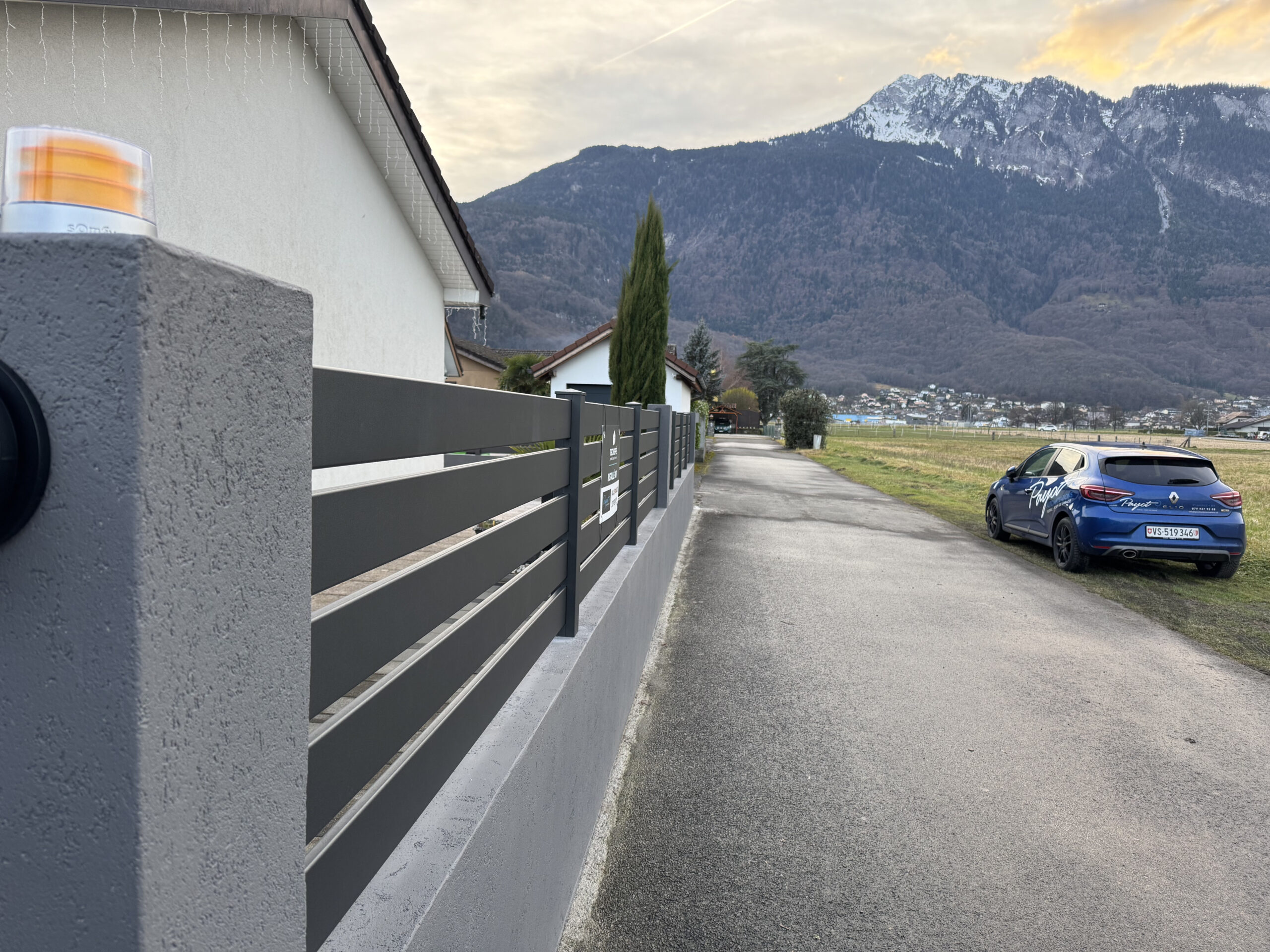 Clôture métallique grise moderne installée, portail extérieur Suisse, rue résidentielle, montagne, voiture bleue.